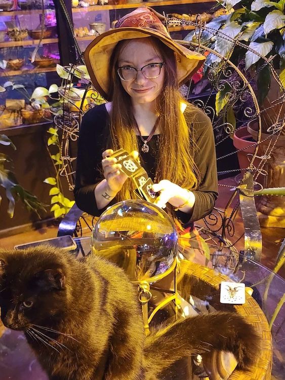 Krista L in witchy attire at a glass table reading a deck of tarot cards